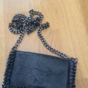 Svart axelväska med kedjerem - Säljer en svart axelväska i skinn med grov kedjerem och dekorativa kedjedetaljer runt kanterna. Väskan har magnetlås och en stilren, lite edgy look. Perfekt för dig som gillar att gömma trender. Den har en liten vit fläck på insidan av skinnet som man ser på bilden men inget som synd alls när väskan är stängd❤️köpt i en butik i Italien och väskan är i äkta skinn❤️