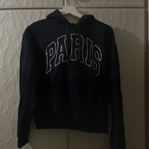 Mörkblå hoodie med PARIS-tryck från H&M - Säljer en mörkblå hoodie från H&M med stort vitt PARIS-tryck på bröstet. Tröjan har huva med dragsko och en klassisk känguruficka framtill. Perfekt för en avslappnad och cool stil. Passar xs-m