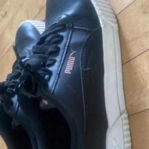 Svarta sneakers från Puma - Svarta Puma sneakers med vit sula och klassisk logga i silver på sidan. Skorna har snörning och är tillverkade i syntetmaterial med SOFTFOAM innersula för extra komfort. Perfekta för en sportig och avslappnad stil.
