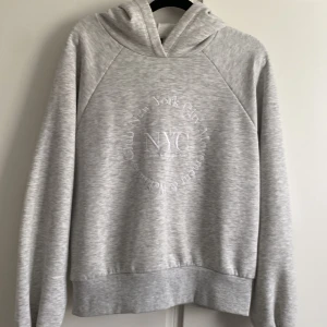 Ljusgrå hoodie med NYC-tryck från Gina Tricot - Säljer en ljusgrå hoodie från Gina Tricot med broderat NYC-tryck och texten 'Wellness Club New York City Movement' på bröstet. Tröjan har huva och långa ärmar, perfekt för en avslappnad stil.