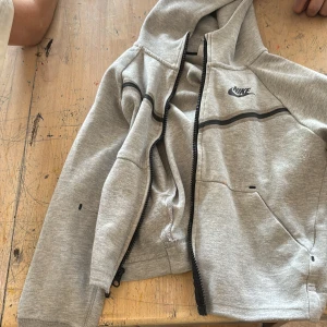 Grå hoodiejacka från Nike - Säljer en grå hoodiejacka från Nike med svart dragkedja och svart Nike-logga på bröstet. Jackan har huva, långa ärmar och en svart linje som detalj över bröstet. Perfekt för träning eller chill.