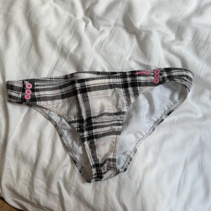 Rutig bikini från Billabong - Säljer en snygg bikini från Billabong i svart, vit och rosa rutig design. Toppen har rosa knappar i mitten och avtagbara axelband, medan trosan har matchande knappar på sidan. Perfekt för sommaren!