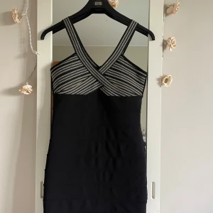 Svart kortklänning med glittriga detaljer från Amisu - Säljer en svart kortklänning från Amisu med glittriga ränder i guld  på axelbanden och överdelen. Klänningen har omlott-detalj upptill och en tight passform som framhäver figuren. Perfekt för dig som vill sticka ut med stil.