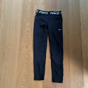 Svarta träningstights från Nike Pro - Säljer ett par svarta Nike Pro tights med hög midja och vit logga på benet. Tightsen är i stretchigt material och har en bred resår i midjan med Nike Pro-tryck. Perfekta för träning eller sport.
