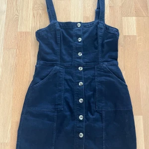 Svart jeansklänning med knappar från Divided - Svart jeansklänning från Divided med justerbara axelband och stora fickor framtill. Klänningen har knäppning med silvriga knappar hela vägen framtill och en tight passform. Perfekt att styla med en t-shirt eller linne under