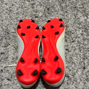 Vita Adidas Copa fotbollsskor - Säljer ett par vita Adidas Copa fotbollsskor med svarta och röda detaljer. Skorna har snörning och är designade för spel på gräs med fasta dobbar. Perfekta för dig som vill ha stilrena och bekväma skor på planen.
