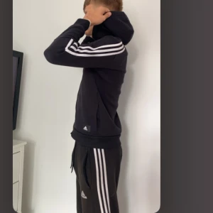 Svart Adidas hoodie med dragkedja och byxor (adidas set) och vita ränder - Säljer ett svart adidas set. Hoodie med dragkedja och klassiska vita ränder längs ärmarna och byxorna. Tröjan har huva, fickor och ribbade muddar. Perfekt för sport eller chill. 