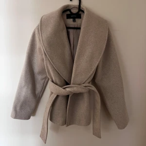 Beige kappa med bälte från Vero Moda - Säljer den populära stilrena beigea kappan från Vero Moda med bred krage och knytskärp i midjan. Jackan har lång ärm och är tillverkad i ett mjukt ullblandat material. Perfekt för höst och vår. Knappt använd.
