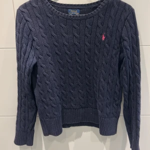 Mörkblå kabelstickad tröja från Polo Ralph Lauren - Säljer en klassisk mörkblå kabelstickad tröja från Polo Ralph Lauren med rund halsringning och röd broderad logga på bröstet. Tröjan har långa ärmar och ribbade muddar vid ärmslut och nederkant. Perfekt för lager-på-lager-stil! Storlek 12-14 år