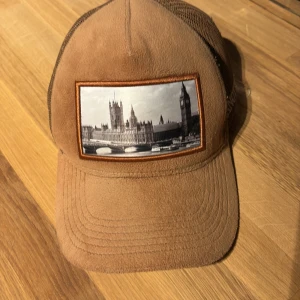 Brun keps från Pianotti med Londonmotiv - Unik brun keps från Pianotti med mesh baktill och justerbar knäppning. Framtill finns ett tygmärke med svartvitt motiv av London och Big Ben. Märkesdetaljer på insidan och baksidan. Kepsen har bra skick 7/10 och mitt pris 300 ( ny pris 800 ). Hör av er vid frågor och funderingar