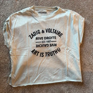 Vit t-shirt från Zadig & Voltaire med tryck - Vit t-shirt från Zadig & Voltaire med svart tryck framtill: 'Rive Droite Est. 1997 Art is Truth' och en liten döskalle. T-shirten har en avslappnad passform och korta ärmar. Perfekt för en cool och casual stil.