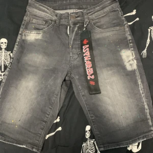 Grå jeansshorts från Dsquared2 - Säljer ett par grå jeansshorts från Dsquared2 med slitna detaljer och färgstänk. Shortsen har fem fickor, bälteshällor och en röd Dsquared2-logga på bakfickan. Nyckelband med Dsquared2 medföljer.