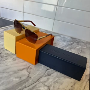 Louis Vuitton solglasögon med guldiga detaljer - Snygga solglasögon från Louis Vuitton med guldiga bågar och bruna skalmar. Glasen är mörktonade och har en fyrkantig form. Märkeslogga på sidan och exklusiv känsla i designen. Perfekt accessoar för att lyfta din stil. Använda en sommar och köpta när jag Bode i Paris.