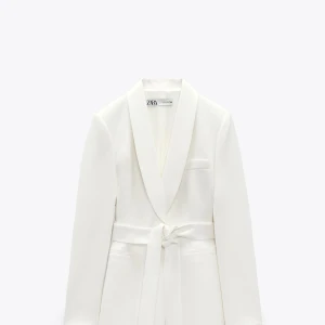 Vit kavaj med bälte från Zara - Stilren vit kavaj från Zara med sjalkrage och knytbälte i midjan. Perfekt för en dressad look och enkel att matcha med olika outfits. Storlek L. Använd 1 gång på studenten. Lite noppigt vid bältet men syns inte då bältet täcker. 