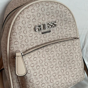 Beige ryggsäck från Guess - Ryggsäck från Guess med justerbara axelremmar och ett litet fack framtill. Väskan har ett allover-mönster med Guess-loggan. Väskan är helt oanvänd, har inga skador eller repor.  Originalpris: ca 1500 kr