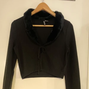 Svart croppad kofta med fuskpäls och knyt - Säljer en svart croppad kofta med mjuk fuskpäls runt kragen och knyt framtill. Tröjan är ribbad och har långa ärmar. Perfekt för att lyfta en outfit med lite extra stil.