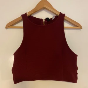 Vinröd croppad linnetopp med dragkedja - Säljer en vinröd croppad linnetopp med breda axelband och rund halsringning. Toppen har en synlig guldfärgad dragkedja bak och är tillverkad i ett ribbat material. Perfekt för att lyfta en outfit!