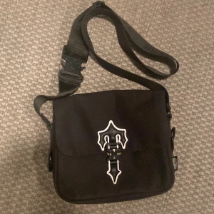 Svart axelväska med vit detalj - Snygg svart axelväska från trapstar med justerbar rem och coolt vitt emblem på framsidan. Väskan har dragkedja på baksidan och praktiskt spänne på axelremmen. Perfekt för att bära dina viktigaste grejer, inte äkta men ser väldigt äkta ut 