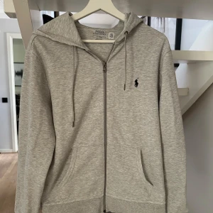 Grå zip hoodiefrån Polo Ralph Lauren - Säljer en stilren grå hoodie från Polo Ralph Lauren i storlek M. Tröjan har dragkedja, huva med dragsko och två fickor framtill. Klassisk logga broderad på bröstet. Perfekt för en avslappnad och snygg look.Nästan aldrig använd. Skriv vid minsta funderingar💯