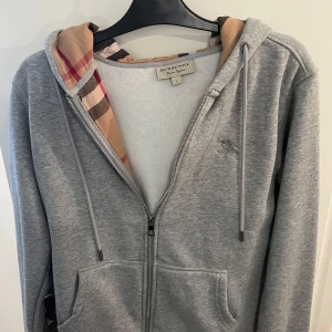 Grå zip hoodie från Burberry - Säljer en grå hoodie från Burberry i storlek L. Tröjan har dragkedja, huva med klassiskt rutigt foder och broderad logga på bröstet. Tillverkad i mjuk bomull för en bekväm känsla. Perfekt för en stilren och avslappnad look. Fick som present men var ej rätt storlek. Kan passa M-L. Tveka inte på att höra av dig vid minsta fråga!