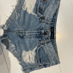 Ljusblå jeansshorts med fransar och slitningar - Säljer ett par ljusblå jeansshorts från One Teaspoon med råa kanter, fransar och stora slitningar både fram och bak. Klassisk femficksmodell med knappgylf och bälteshällor. Perfekta för en avslappnad stil.