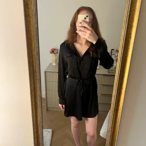 Svart skjortklänning med knytband - Säljer en svart skjortklänning med lång ärm och knytband i midjan. Klänningen har krage, knappar framtill och två bröstfickor. Perfekt till alla årstider.