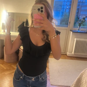 Svart omlott bodysuit med volang - Säljer en svart bodysuit med djup v-ringning och bred volang längs urringningen. Modellen har breda axelband och är åtsittande. Perfekt för dig som vill ha en stilren och trendig topp till jeans eller kjol. PRISET KAN DISKUTERAS🩷🩷