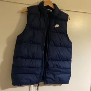 Mörkblå dunväst från Nike - Snygg mörkblå dunväst från Nike med hög krage och dragkedja framtill. Västen har en klassisk puffer-look och vit Nike-logga på bröstet. Perfekt för lager-på-lager och enkel att matcha med olika outfits.
