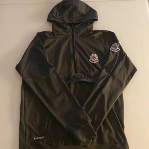 Mörk grön vindjacka med huva från Moncler - Säljer en svart vindjacka från Moncler med huva och halv dragkedja framtill. Jackan har Moncler-logga på bröstet och ärmen samt en diskret logotyp nertill. Medföljer svarta snören till huvan.