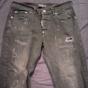 Svarta jeansbyxor från Dsquared2 - Säljer ett par svarta jeansbyxor från Dsquared2 med slitningar och unika detaljer framtill. Byxorna har klassisk femficksdesign, silverfärgade knappar och en rak passform. Perfekta för dig som vill ha en cool och trendig look.