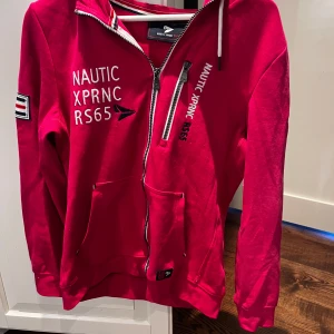 Röd hoodie från Nautic XPRNC RS65 - Röd hoodie med dragkedja från Nautic XPRNC RS65. Har vita tryckdetaljer, bröstficka med dragkedja och fickor framtill. Tillverkad i ekologisk bomull och har huva med snörning samt randiga detaljer vid dragkedjan.