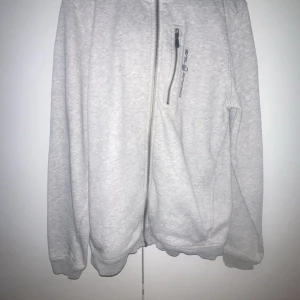 Ljusgrå hoodiejacka med dragkedja - Ljusgrå hoodiejacka med hel dragkedja framtill och en bröstficka med dragkedja. Hoodien har huva och ribbade muddar vid ärmslut och nederkant. Perfekt för en avslappnad stil. Hör av er vid funderingar😁