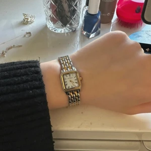 Mockberg klocka i guld och silver - Säljer en snygg Mockberg klocka med fyrkantig vit urtavla och romerska siffror. Armbandet är i både guld och silverfärg, vilket ger en stilren och klassisk look. Perfekt accessoar för att lyfta din outfit.