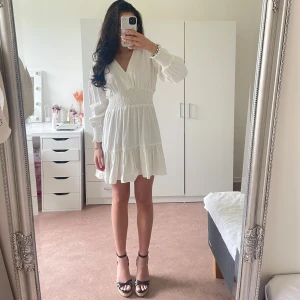 Vit kortklänning med v-ringning och volang - Superfin vit klänning med v-ringad hals, långa ärmar och smock i midjan. Klänningen har volangdetalj nertill och är i ett lätt, luftigt material. ENDAST ANVÄND 1 GÅNG. Perfekt till student/skolavslutning/konfirmation/midsommar osv🌸Storlek Xs men passar även xxs-xs-s då den är stretchig i midjan☺️