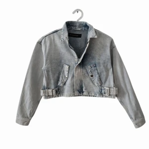 Croppad jeansjacka från Never Denim - Snygg croppad jeansjacka från Never Denim i ljusblå denim. Jackan har klassisk krage, dragkedja framtill, två fickor med lock och coola spänndetaljer i midjan. Perfekt för dig som vill ha en trendig och avslappnad look.