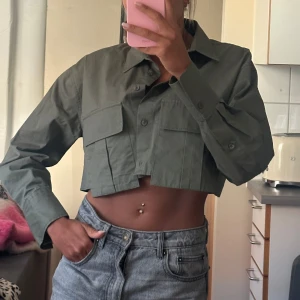 Croppad grön skjorta från Zara - Snygg croppad skjorta från Zara i militärgrönt. Skjortan har stora bröstfickor, klassisk krage och knäppning framtill. Tillverkad i bomull och har långa ärmar. 