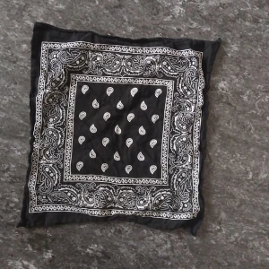 Svartvit paisley bandana halsduk - Snygg svart halsduk i bandana-stil med klassiskt vitt paisleymönster. Fyrkantig form, tunn och lätt att styla runt halsen eller i håret. Perfekt accessoar för att ge din outfit en cool touch.