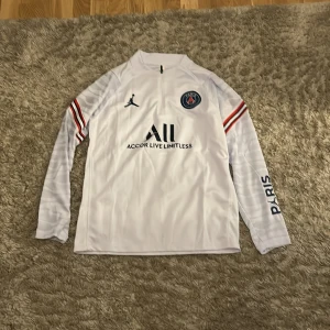 Vit PSG långärmad fotbollströja Jordan - Snygg vit långärmad PSG-tröja från Jordan-kollektionen. Tröjan har svarta och röda detaljer på ärmarna, PSG-emblem på bröstet och tryck med 'PARIS' och 'VISIT RWANDA'. Tillverkad i lätt och ventilerande funktionsmaterial. Tröjan passar i storlek 158cm/164cm