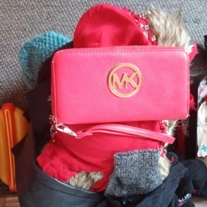 Röd Michael Kors plånbok i skinn - Snygg röd plånbok från Michael Kors med stort guldfärgat MK-emblem framtill. Plånboken är i skinn och har en avtagbar handledsrem, perfekt för att bära med sig mobilen och korten. Stilren och trendig accessoar för dig som gillar färg.