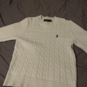 Vit kabelstickad tröja Polo Ralph Lauren - En vit och mycket snygg och välbehandlad kabelstickad tröja från Polo Ralph Lauren med klassisk rund halsringning och broderad logga på bröstet. Tröjan har ribbade muddar och är långärmad, perfekt för lager-på-lager under kyliga dagar. Står fel på tröjan är faktiskt i M storlek