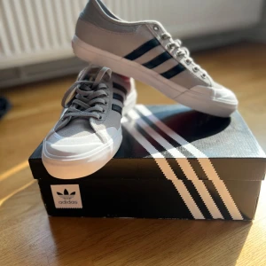 Adidas Matchcourt grå sneakers - Snygga Adidas Matchcourt sneakers i grå canvas med klassiska svarta ränder på sidorna och vit tå. Skorna har röda yttersulor och vita detaljer runt sulan. Perfekta för dig som gillar streetstyle och vill ha en clean look med retrovibbar. Aldrig använda 