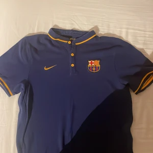 Blå FC Barcelona Nike fotbollströja - Snygg blå FC Barcelona fotbollströja från Nike med klubbmärke på bröstet och gula detaljer på krage, knappar och ärmslut. Klassisk pikékrage och korta ärmar. Perfekt för dig som vill visa vilket lag du håller på.