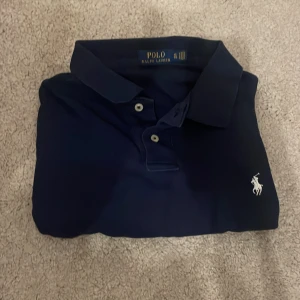 Marinblå pikétröja Polo Ralph Lauren XL - Klassisk marinblå pikétröja från Polo Ralph Lauren i storlek XL. Tröjan har vit broderad logga på bröstet, krage och två knappar framtill. Perfekt för en clean och stilren look. Tillverkad i mjuk bomull som känns skön mot huden.