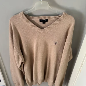 Beige stickad tröja från Gant - Beige stickad tröja från Gant med v-ringad hals och diskret broderad logga på bröstet. Tröjan har långa ärmar och ribbade muddar vid ärmslut och nederkant. Perfekt för en chill och stilren look.