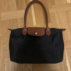 Svart Longchamp handväska med skinn - Klassisk svart handväska från Longchamp med bruna skinnhandtag och lock. Väskan har guldfärgad dragkedja med Longchamp-logga och detaljer i präglat skinn. Perfekt storlek för vardagens essentials och stilren look.