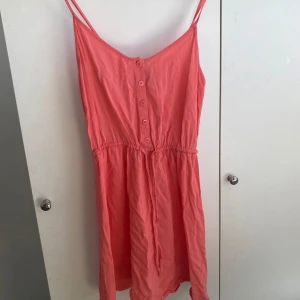 Korallrosa sommarklänning med knappar - Säljer en söt korallrosa klänning med smala axelband och knappar framtill. Klänningen har en markerad midja och är tillverkad i ett lätt bomullsmaterial, perfekt för varma dagar.