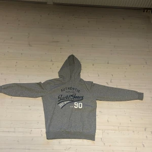Grå hoodie från Jack & Jones - Snygg grå hoodie från Jack & Jones med tryck i mörkblått och vitt på bröstet. Klassisk huva och långa ärmar, perfekt för en chill och avslappnad stil. Mjuk bomullsblandning som känns skön mot huden. 