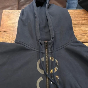 Mörkblå hoodie från Hugo med dragkedja - Snygg mörkblå hoodie från Hugo med stor huva, hel dragkedja och tryckt logga i silver på framsidan. Tillverkad i mjuk bomullsmix och fleecefodrad på insidan för extra komfort. Perfekt för dig som gillar stilrena och bekväma plagg. Pris kan diskuteras!