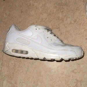 Nike Air Max 90 helvita sneakers - Klassiska Nike Air Max 90 sneakers i helvitt skinn och syntet. Snygg chunky sula med synlig Air Max-bubbla och diskreta detaljer. Perfekta för dig som gillar en clean och sportig look. Ikonisk Nike-logga på sidan och bak.