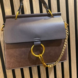 Stilren handväska med guldkedja - Snygg handväska i skinn med brunt lock och detaljer i guld. Väskan har en kedja och stor guldring framtill, samt handtag upptill. Finns i tre färgkombinationer: mörkbrun/svart, mörkbrun/ljusbrun och mörkbrun/beige. Perfekt för dig som gillar klassisk men trendig stil. Väskan finns i 3 färger svart, brun, rosa